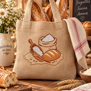 Può includere: Una borsa in juta color sabbia con un ricamo raffigurante un sacco di farina, un cucchiaio di legno e una pagnotta. La borsa contiene due lunghe baguette croccanti e ha un asciugamano in lino a righe beige e rosse drappeggiato sul lato.