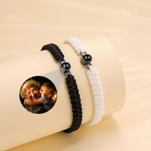 Könnte beinhalten: Zwei geflochtene Armbänder, eines in Schwarz und eines in Weiß, mit jeweils einem runden, dunkelmetallenen Anhänger in der Mitte. Das schwarze Armband projiziert einen kreisförmigen Lichtstrahl mit einem Farbfoto einer älteren Person und eines Kindes.