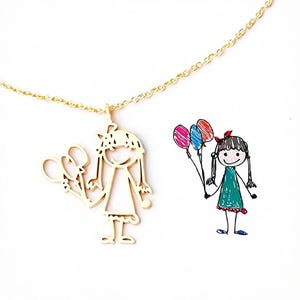 Op de afbeelding: Een goudkleurige metalen ketting met een hanger in de vorm van een kindertekening van een persoon die drie ballonnen vasthoudt. Naast de hanger staat de originele gekleurde tekening van een meisje in een groene jurk met rode en blauwe ballonnen.