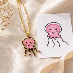 Op de afbeelding: Een goudkleurige metalen ketting met een hanger in de vorm van een lachende roze kwal met zwarte omlijningen. De hanger wordt getoond naast een witte kaart met een handgetekende illustratie van hetzelfde roze kwalfiguurtje.