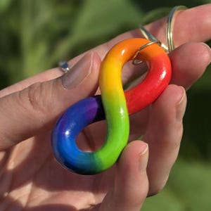 Puede incluir: Un llavero con forma de símbolo de infinito liso y brillante, con un degradado de arcoíris vibrante en rojo, naranja, amarillo, verde, azul y morado. El diseño curvo y continuo está unido a una anilla de metal dorado para llaves.