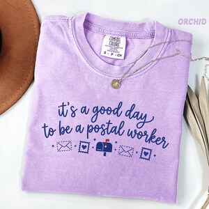 Könnte beinhalten: Ein hellviolettes Kurzarm-T-Shirt mit Rundhalsausschnitt. Das Shirt zeigt einen dunkelblauen Schriftzug, der lautet, it's a good day to be a postal worker, begleitet von kleinen grafischen Symbolen von Briefumschlägen, Briefmarken und einem Briefkasten.