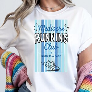 May include: A white t-shirt featuring a graphic design with vertical light and dark blue stripes. The text reads Mediocre RUNNING Club and SLOW IS A PACE, accompanied by a small line-art illustration of a running shoe and four-pointed stars.