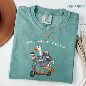 以下が含まれることがあります： ライトグリーンのクルーネックTシャツ。ブレザーとネクタイを着用し、コーヒーカップを持ってバックパックを背負い、赤いスクーターに乗っているガチョウのグラフィックが描かれています。ガチョウのイラストの上には「off to update the timeline」という文字がプリントされています。