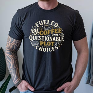 Könnte beinhalten: Ein anthrazitfarbenes Kurzarm-T-Shirt mit einer kreisförmigen Grafik und dem Text FUELED BY COFFEE AND QUESTIONABLE PLOT CHOICES. Das Design enthält Illustrationen einer Kaffeetasse, eines offenen Buches, einer Feder und verstreuter Kaffeebohnen.