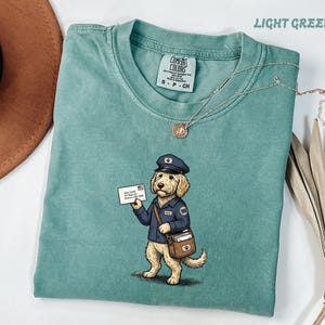 Könnte beinhalten: Ein hellgrünes Kurzarm-T-Shirt mit einer Grafik eines Golden Retrievers als Postbote, der eine blaue Uniform, eine Mütze und eine Umhängetasche trägt und einen Umschlag mit dem Text John Smith 123 Maple St Anytown, USA 12345 hält.