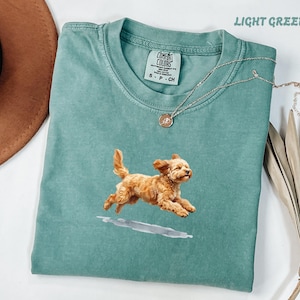 Puede incluir: Una camiseta de manga corta de color verde claro con el gráfico de un perro de color marrón dorado saltando sobre un pequeño charco. La camiseta tiene una etiqueta de la marca Comfort Colors en el cuello y un collar de cadena plateada.