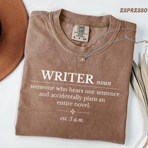 Może przedstawiać: Brązowy t-shirt z krótkim rękawem i okrągłym dekoltem, z białym nadrukiem: WRITER noun someone who hears one sentence and accidentally plans an entire novel. est. 3 a.m. Koszulka posiada metkę z oznaczeniem marki Comfort Colors.