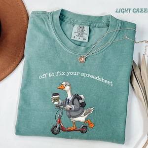 Op de afbeelding: Een lichtgroen t-shirt met een afbeelding van een gans met een bril, een colbert en een rugzak, rijdend op een rode step en met een koffiebeker in de hand. De tekst "off to fix your spreadsheet" staat in witte letters boven de gans.