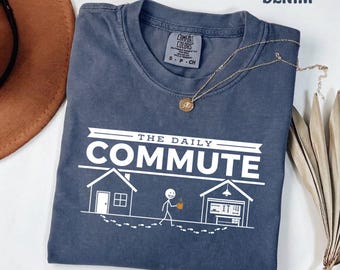 May include: A denim blue short-sleeve t-shirt featuring a white graphic print that reads THE DAILY COMMUTE. The design includes a house, a stick figure holding a mug, and a garage with tools, connected by a path of footprints. The shirt has a crew neck.