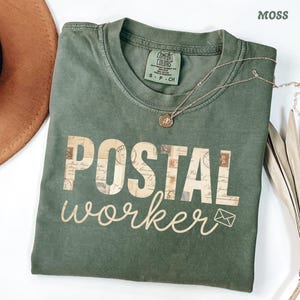 Könnte beinhalten: Ein moosgrünes Kurzarm-T-Shirt mit Rundhalsausschnitt. Auf der Vorderseite steht der Text POSTAL in Blockbuchstaben, die mit einem Vintage-Briefmarkenmuster gefüllt sind, und das Wort worker in Schreibschrift, gefolgt von einem Briefumschlag.