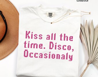 May include: A white short-sleeved t-shirt featuring the pink pixelated text "Kiss all the time. Disco, Occasionaly" printed on the front. The shirt has a crew neckline and a Comfort Colors brand label visible at the inside of the collar.