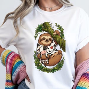 Könnte beinhalten: Ein weißes T-Shirt mit einer grafischen Illustration eines braunen Faultiers, das an einem moosigen Ast hängt. Das Faultier trägt ein weißes Oberteil mit schwarzem Kuhmuster und dem Text I heart COWS und hält ein kleines schwarz-weißes Kuscheltier.