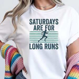 May include: A white short-sleeved t-shirt featuring the dark teal text "SATURDAYS ARE FOR LONG RUNS" and a central graphic of a runner in silhouette against a background of horizontal light teal stripes with a distressed texture.