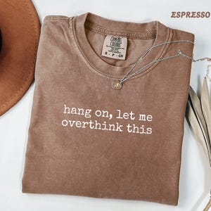 May include: An espresso-coloured short-sleeve t-shirt with a crew neckline. The shirt features the white text "hang on, let me overthink this" printed on the front. A silver-toned necklace with a small circular pendant rests on the shirt.