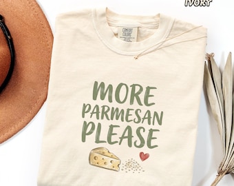 May include: An ivory crewneck t-shirt featuring the green text MORE PARMESAN PLEASE, a graphic of a wedge of Swiss cheese, and a small red heart. The shirt has a Comfort Colors brand label at the inner neckline and is labeled as ivory color.