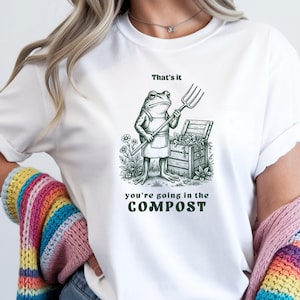 May include: A white t-shirt featuring a dark green graphic of a frog wearing an apron and holding a pitchfork next to a wooden compost bin. The text above the frog reads That's it and the text below reads you're going in the COMPOST.