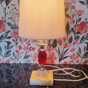 May include: A table lamp with a cream-coloured, vertically ribbed cylindrical shade featuring a bright green scalloped trim. The lamp base consists of a square, marbled beige stone block, a faceted clear crystal accent, and a red glass centre.