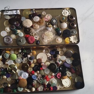 May include: Two open vintage metal tins filled with a variety of buttons. The buttons are in various colours, sizes, and materials, including plastic, metal, and fabric. Some buttons are round, others are square, and some have unique shapes. The tins are on a light-coloured surface.