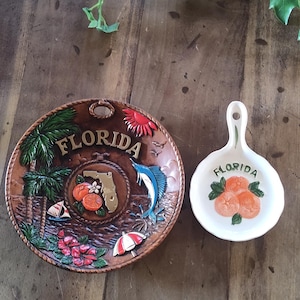May include: A round brown decorative plate featuring a raised Florida map, palm trees, a marlin, and citrus fruit, alongside a small white ceramic spoon rest with a painted orange design and the word Florida printed in green lettering.