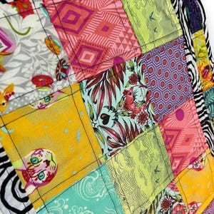 May include: Close-up of a colorful patchwork textile. The design features squares with various patterns, including geometric shapes, floral motifs, and animal illustrations. The color palette includes yellow, pink, green, and purple, with black stitching outlining each square.
