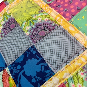 May include: Close-up of a colorful patchwork quilt with a variety of floral and geometric patterns. The quilt features squares of blue, green, purple, and pink fabrics, stitched together with red thread. The quilt is a mix of textures and colors.