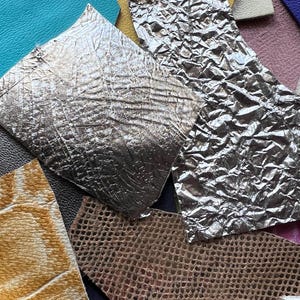 Op de afbeelding: Een verzameling diverse leer- en kunstleerstalen in verschillende kleuren en texturen, waaronder metallic zilver, turquoise, geel, paars, roze en bruin met reliëfpatronen, reptielachtige schubben en gladde afwerkingen.