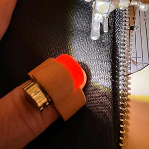 Könnte beinhalten: Ein am Finger befestigtes LED-Nählicht mit beigefarbenem Kunststoffgehäuse und hellem orangefarbenem Leuchten, positioniert über schwarzem Stoff neben einem Nähmaschinen-Nähfuß und einer silbernen Stichplatte mit Messmarkierungen.