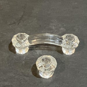 May include: A clear glass cabinet handle with a curved center bar and two faceted, circular mounting ends, accompanied by a single matching faceted, circular cabinet knob. Both pieces feature a transparent, reflective finish and geometric detailing.