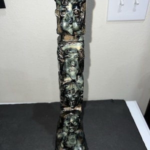 May include: A tall, vertical ceramic sculpture featuring a stack of four distinct, expressive faces carved in a dark, mottled green and black glaze. The figures are arranged in a column, each with unique facial features and textured details.