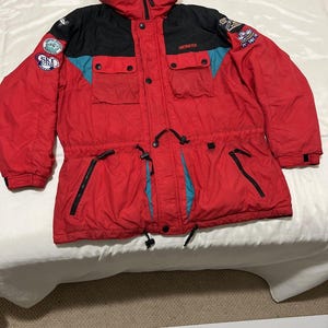 May include: A red, black, and teal winter jacket with a hood, featuring the Obermeyer brand name on the chest, multiple zippered pockets, and various circular patches on the sleeves including Ski Utah and Crystal Mountain Washington logos.