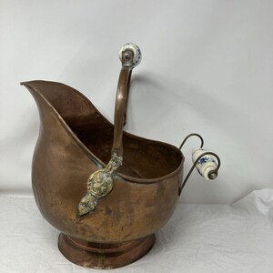 May include: A copper coal scuttle with a wide, open-top basin, a pedestal base, and a tall, arched handle. The handle and the smaller rear handle feature white ceramic grips decorated with blue floral patterns and brass-toned metal accents.