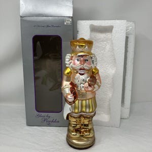 May include: A 10-inch glass nutcracker figurine featuring a gold crown, white beard, and gold-striped trousers. The figure is painted in shades of gold, white, and peach, and is displayed in front of its original silver box labeled Glass by Peschka.