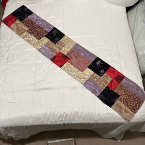 May include: A rectangular patchwork table runner with a variety of fabrics in red, black, gold, brown, and purple. The runner is made of squares of different patterns and textures, creating a unique and decorative piece.