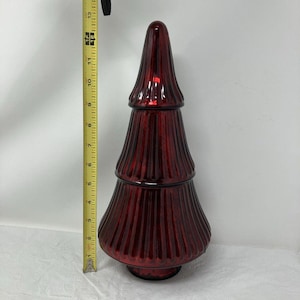 May include: A deep red, ribbed glass tree-shaped decorative object. A yellow measuring tape placed vertically next to the object indicates a total height of approximately 15 inches from the base to the pointed top of the glass structure.