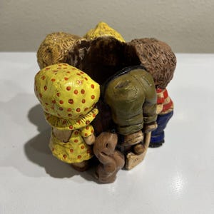 May include: A circular resin planter featuring a group of children and a small dog standing around the exterior. The children wear colorful clothing including a yellow polka-dot dress, olive green pants, and a red and white striped shirt.