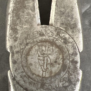 May include: A pair of silver-toned metal pliers featuring a circular embossed logo in the center. The logo depicts a person climbing a pole and includes the text M. KLEIN & SONS and CHICAGO U.S.A. The metal surface shows signs of wear and texture.
