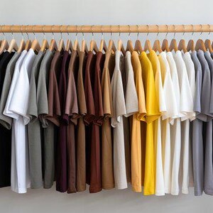 May include: A collection of short-sleeved t-shirts hanging on wooden hangers from a wooden rod. The shirts are arranged in a gradient color spectrum, ranging from black and gray to white, brown, tan, yellow, and various shades of blue and gray.