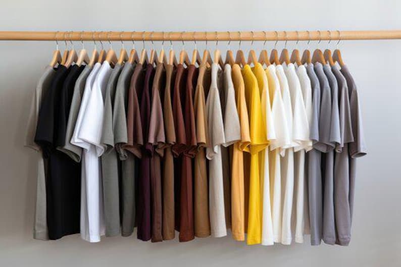 May include: A collection of short-sleeved t-shirts hanging on wooden hangers from a wooden rod. The shirts are arranged in a gradient color spectrum, ranging from black and gray to white, brown, tan, yellow, and various shades of blue and gray.