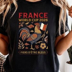 May include: A black t-shirt featuring a graphic of a rooster holding a French flag, a soccer ball, and decorative foliage. The text reads "FRANCE WORLD CUP 2026" at the top and "FIERS D'&Ecirc;TRE BLEUS" at the bottom in a rectangular frame.