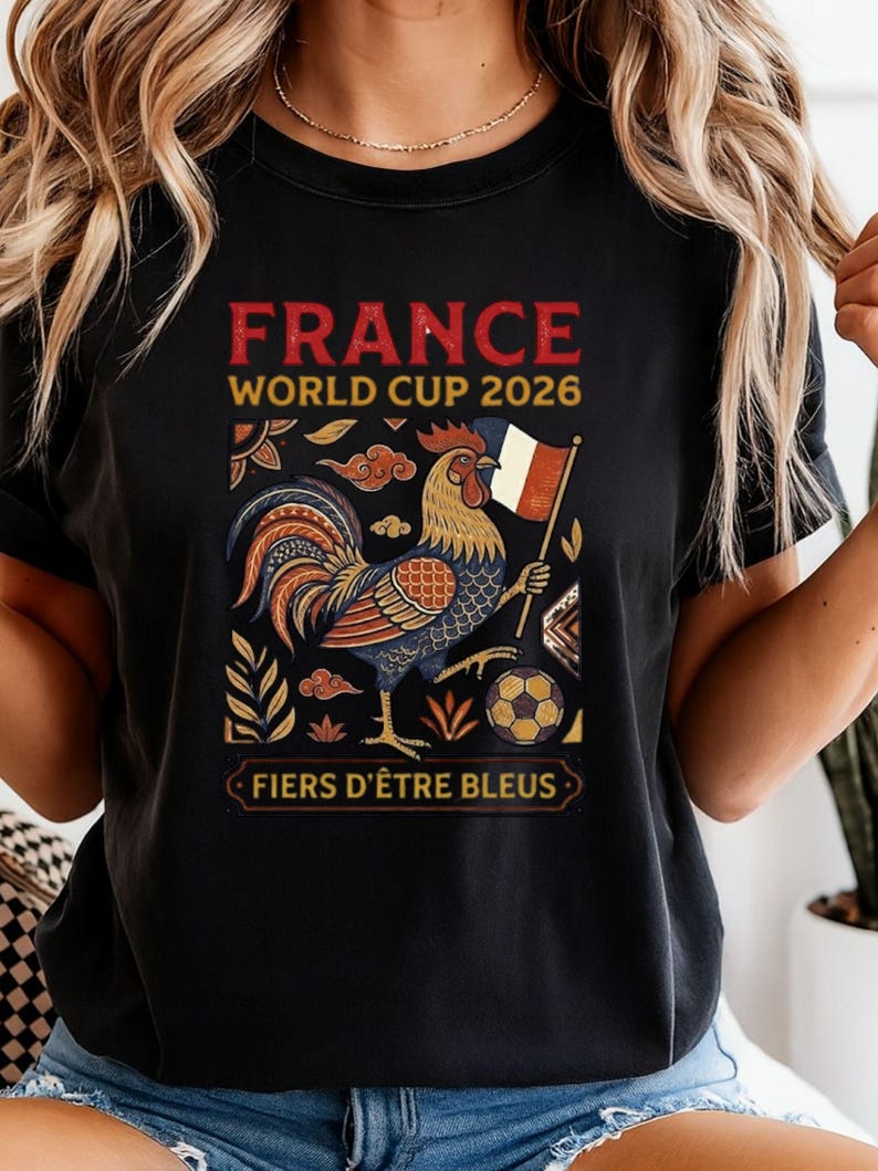 May include: A black t-shirt featuring a graphic of a rooster holding a French flag, a soccer ball, and decorative foliage. The text reads "FRANCE WORLD CUP 2026" at the top and "FIERS D'&Ecirc;TRE BLEUS" at the bottom in a rectangular frame.