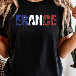 May include: Black t-shirt featuring the word "FRANCE" in the colors of the French flag. The letters are interspersed with silhouettes of soccer players in action, creating a dynamic and patriotic design. A stylish tee for soccer fans.