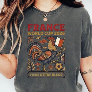 May include: A charcoal gray t-shirt featuring a graphic of a rooster holding a French flag and a soccer ball. The text reads FRANCE WORLD CUP 2026 and FIERS D'&Ecirc;TRE BLEUS. The design includes decorative leaf and cloud patterns in red, blue, and gold.