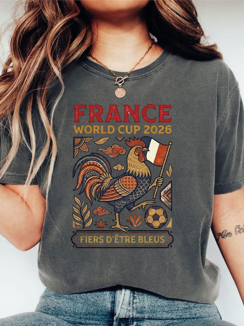 May include: A charcoal gray t-shirt featuring a graphic of a rooster holding a French flag and a soccer ball. The text reads FRANCE WORLD CUP 2026 and FIERS D'&Ecirc;TRE BLEUS. The design includes decorative leaf and cloud patterns in red, blue, and gold.