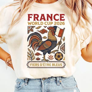 May include: A cream-colored t-shirt featuring a graphic print with the text FRANCE WORLD CUP 2026 and FIERS D'&Ecirc;TRE BLEUS. The design includes a colorful rooster holding a French flag next to a soccer ball, surrounded by decorative leaf and cloud motifs.