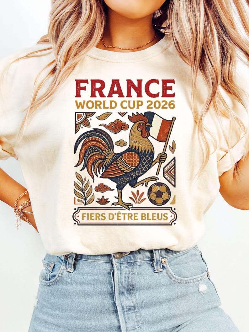 May include: A cream-colored t-shirt featuring a graphic print with the text FRANCE WORLD CUP 2026 and FIERS D'&Ecirc;TRE BLEUS. The design includes a colorful rooster holding a French flag next to a soccer ball, surrounded by decorative leaf and cloud motifs.