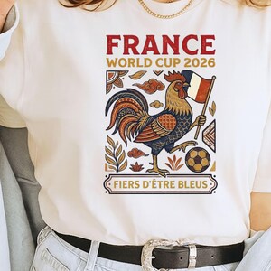 May include: A cream-colored t-shirt featuring a graphic of a rooster holding a French flag and a soccer ball. The text reads "FRANCE WORLD CUP 2026" and "FIERS D'&Ecirc;TRE BLEUS" in red and gold lettering, surrounded by decorative geometric and floral patterns.