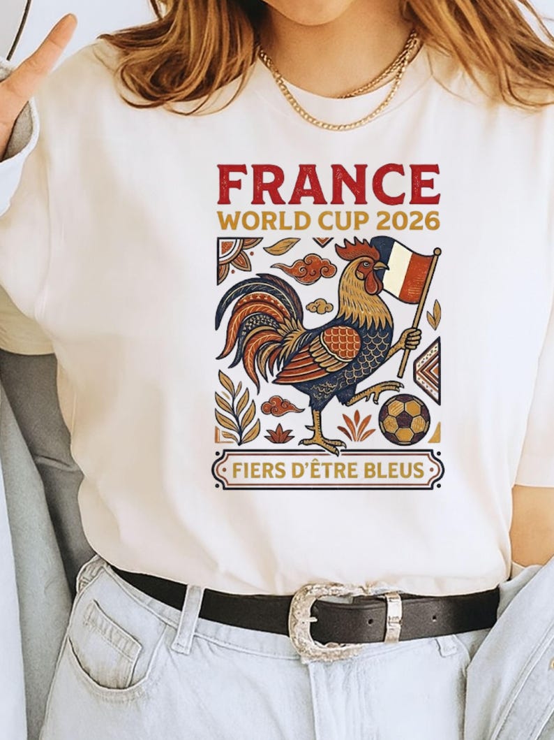 May include: A cream-colored t-shirt featuring a graphic of a rooster holding a French flag and a soccer ball. The text reads "FRANCE WORLD CUP 2026" and "FIERS D'&Ecirc;TRE BLEUS" in red and gold lettering, surrounded by decorative geometric and floral patterns.