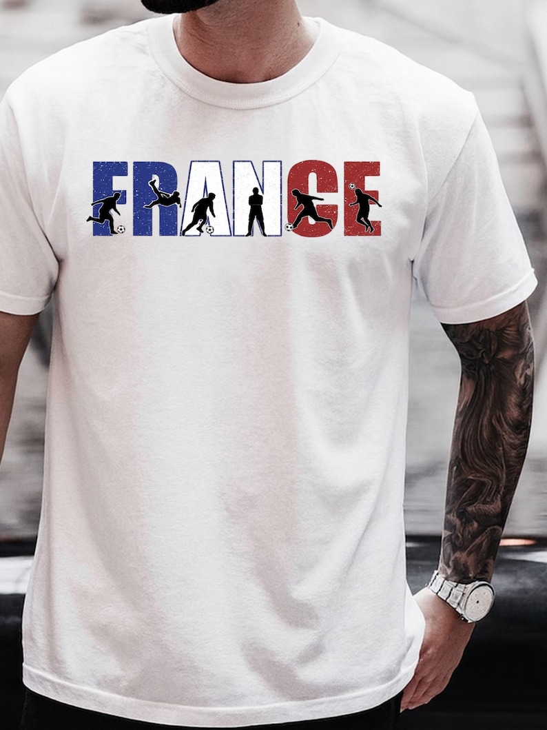 May include: White t-shirt featuring the word "FRANCE" in blue and red, with black silhouettes of soccer players incorporated into the letters. The shirt has a crew neck and short sleeves.
