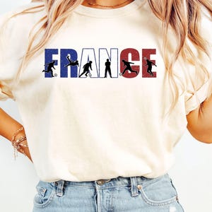 May include: Cream-colored t-shirt featuring the word "FRANCE" in blue, white, and red, with silhouettes of soccer players. The shirt has short sleeves and a crew neck.